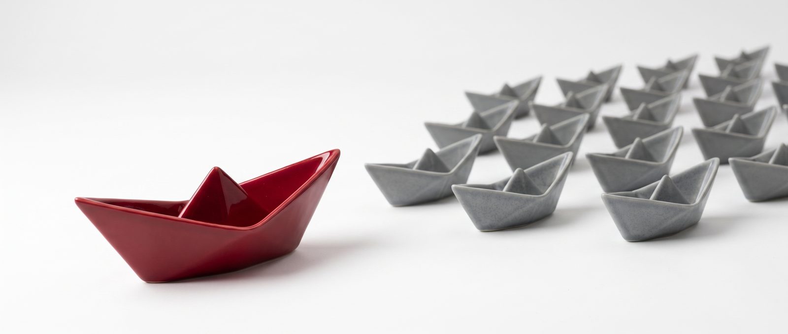 A single glossy cherry-red ceramic paper boat set apart from, and facing, a fleet of identical matte gray ceramic paper boats on a white studio backdrop.